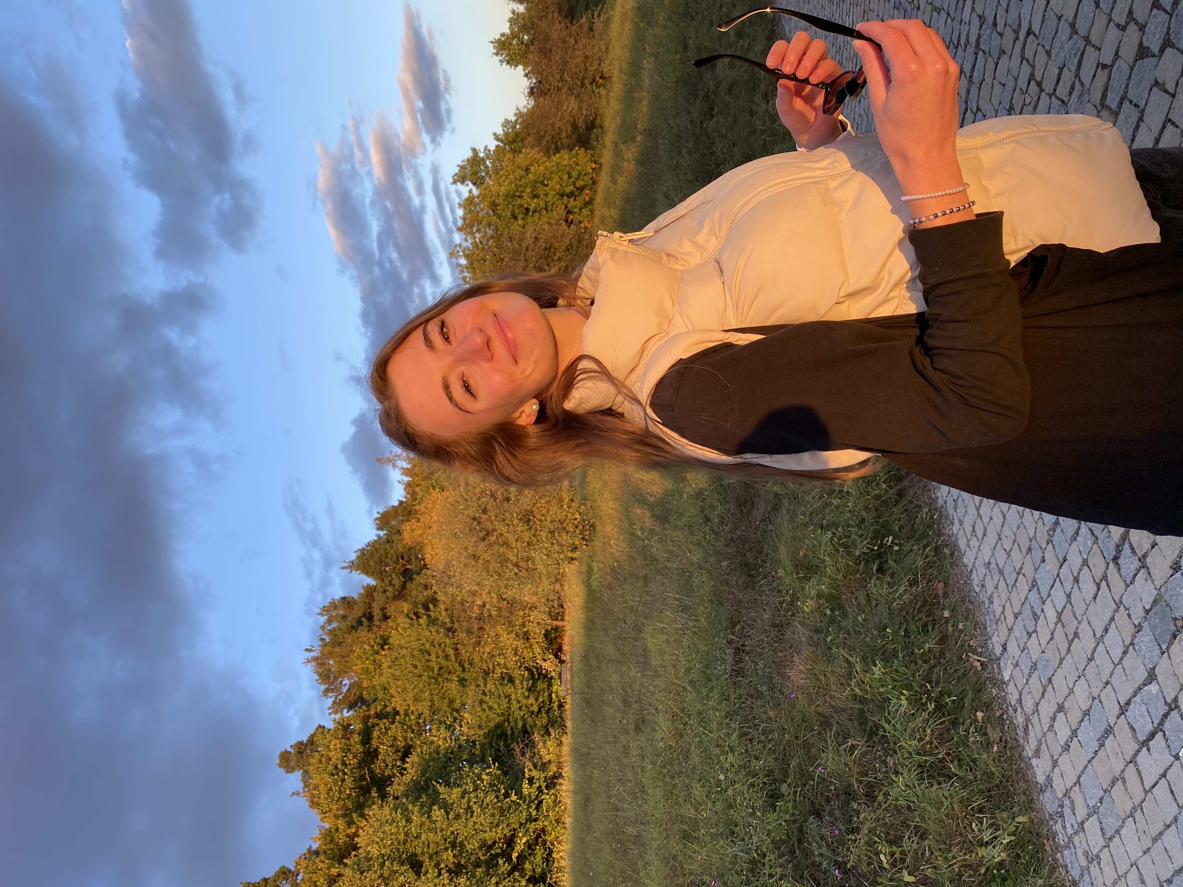 Portrait of a smiling girl with long brown hair wearing black turtleneck and white vest, holding sunglasses during golden hour. Standing in a garden behind trees, with a moody skies.